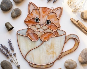Puede incluir: Un atrapasoles de vidrieras que muestra un gato atigrado naranja sentado dentro de una taza de té blanca. El gato tiene segmentos de vidrio detallados con bordes de lámina de cobre, ojos oscuros y patas que descansan sobre el borde de la taza.
