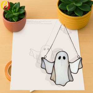 May include: A handcrafted, stained-glass ghost with outstretched arms, hanging from thin wires. The ghost is primarily white with black eyes and a dark outline. The image includes a paper with a ghost outline and two potted succulents.