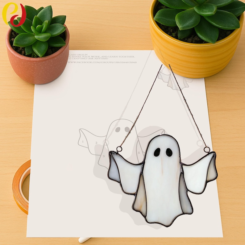 May include: A handcrafted, stained-glass ghost with outstretched arms, hanging from thin wires. The ghost is primarily white with black eyes and a dark outline. The image includes a paper with a ghost outline and two potted succulents.