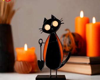 May include: A black cat figurine with orange accents, holding a shovel. The cat has large, round, glowing eyes and is mounted on a black rectangular base. The background includes lit orange candles and a small pumpkin.