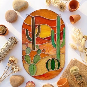 Op de afbeelding: Een ovaal glas-in-loodpaneel met een woestijnlandschap met een gele zon, een oranje lucht en diverse groene cactussen, waaronder saguaro- en vijgcactussen. De glasstukken zijn verbonden door dunne metalen lijnen in een decoratief wandstuk.