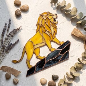 May include: A stained glass lion figure featuring yellow glass panels with copper foil soldering. The lion is depicted in a roaring stance, standing atop a jagged, dark black glass rock formation. The piece is constructed with distinct glass segments.
