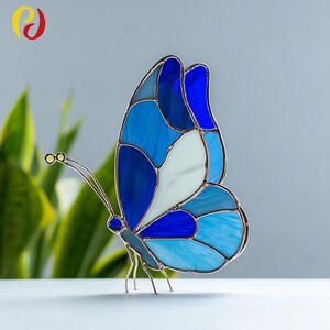 Può includere: Una scultura a farfalla in vetro colorato nei toni del blu e del bianco. La farfalla ha un design dettagliato con una cornice in metallo e delicate antenne. Le ali presentano varie tonalità di blu, creando un pezzo vibrante e accattivante.