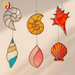 May include: Six stained glass seashell ornaments in various shapes and colours, including white, yellow, orange, pink, and blue. Each piece is outlined in a metallic frame and suspended by a chain, ideal for coastal decor.