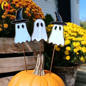 May include: Three Halloween ghost decorations with black witch hats, set on metal stakes. The ghosts are in shades of white and light blue, with black oval eyes. They are placed on top of an orange pumpkin, with yellow and orange flowers in the background.