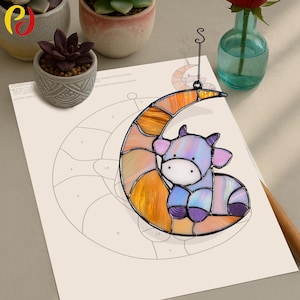 Peut inclure: Un attrape-soleil en verre coloré représentant une vache violette assise sur un croissant de lune. La lune est faite de verre orange et jaune avec une finition texturée. L'attrape-soleil est suspendu à une chaîne en métal noir.