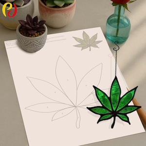 May include: A green stained glass cannabis leaf with black outlines hangs near a drawing of a cannabis leaf. The drawing is on a white sheet of paper. Small potted succulents and a vase with a red rose are in the background.