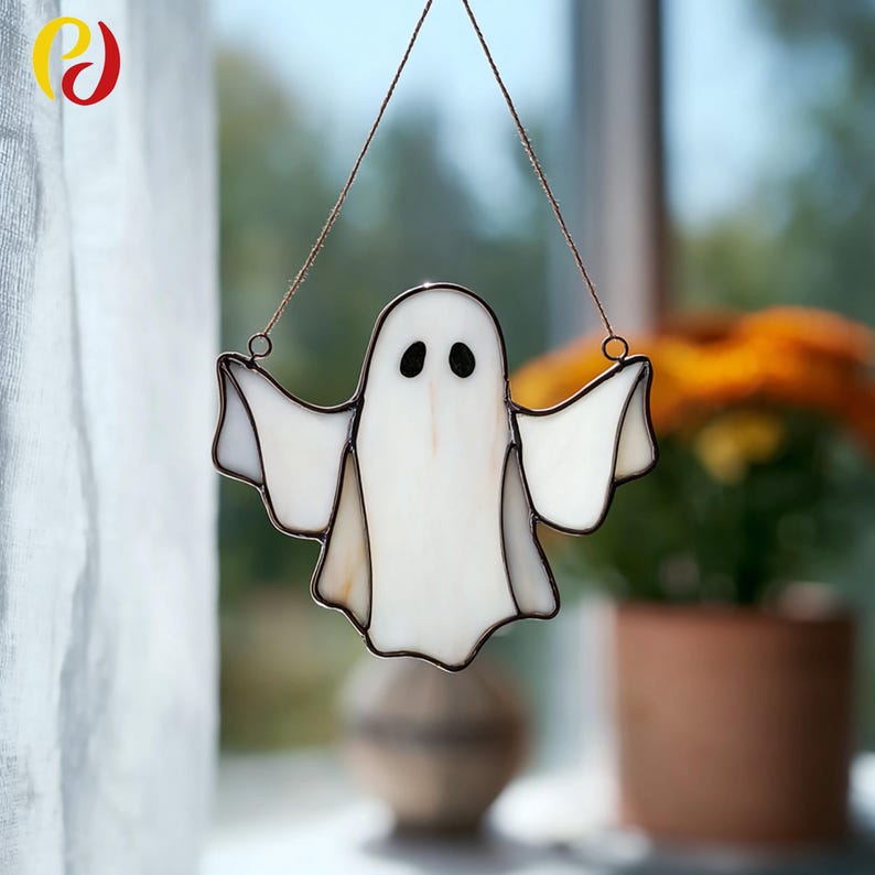 May include: A stained glass ghost decoration with a white body, black eyes, and outstretched arms. The ghost is outlined in a dark brown color and hangs from a thin wire. A Halloween-themed decoration.