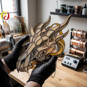 May include: A stained glass dragon skull in cream, grey, and yellow. The artwork is held by gloved hands and features detailed craftsmanship. A decorative chain is attached for hanging. The piece is likely handmade and suitable for home decor.