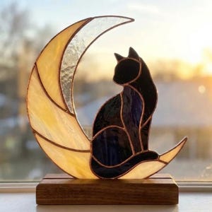 Peut inclure: Un attrape-soleil en vitrail représentant la silhouette d'un chat noir assis sur un croissant de lune en verre texturé jaune pâle et blanc. L'objet est fixé sur une base rectangulaire en bois, avec des lignes de soudure en cuivre.