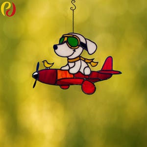 May include: A stained glass suncatcher featuring a white dog wearing goggles and a yellow scarf, flying a red airplane with a yellow bird on the wing.