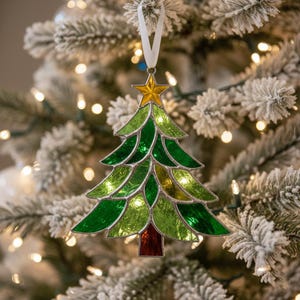 May include: A stained glass Christmas tree decoration with a gold star. The tree is made of green and yellow glass with a red trunk. It hangs from a white ribbon against a snowy tree background.