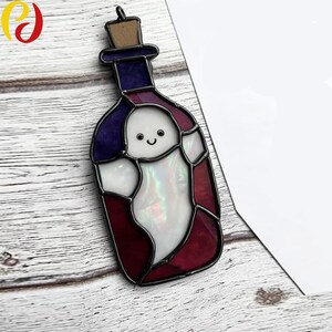Può includere: Un ornamento a forma di bottiglia in vetro colorato con un fantasma sorridente all'interno. La bottiglia ha una parte superiore viola, lati rossi e un fantasma bianco iridescente. Un tappo di sughero completa la bottiglia. L'ornamento è su una superficie di legno.