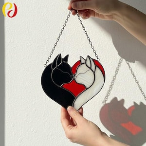 May include: A stained glass heart featuring two cats, one black and one white, kissing. The heart shape has a red centre and is suspended by a chain. The artwork is held up by a person's hands.