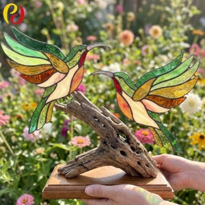 Può includere: Due colibrì in vetro colorato con ali verdi, gialle e ambrate e petto bianco, montati su un pezzo di legno galleggiante fissato a una base rettangolare in legno. Gli uccelli sono posizionati come se stessero volando l'uno verso l'altro.