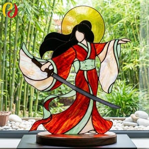 May include: Stained glass sculpture of a woman in a red kimono holding a sword. The figure has black hair and a yellow halo. The sculpture is on a wooden base, with a bamboo background.