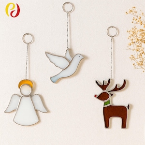 Op de afbeelding: Drie hangende glas-in-lood ornamenten: een engel met een gouden halo, een witte duif en een bruin rendier met een rode neus en groene kraag. Elk ornament is opgehangen aan een zilveren ketting en ring.