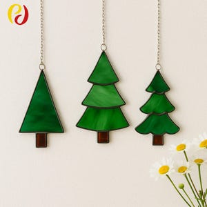 Könnte beinhalten: Drei Weihnachtsbaum-Ornamente aus Glasmalerei. Jeder Baum hat eine andere Form, mit dunkelgrünem Glas und einem braunen Stamm. Die Bäume hängen an silbernen Ketten. Der Hintergrund ist hellbeige.