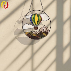 May include: A round stained glass window featuring a colorful hot air balloon, white clouds, and mountain scenery. The window is framed in black metal and hangs from a chain. The balloon has yellow, green, blue, and red sections.