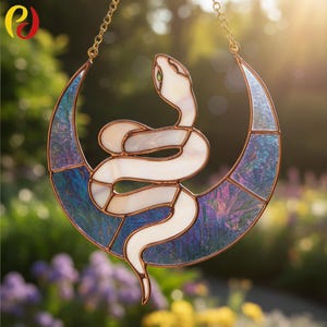 Pode incluir: Um apanhador de sol em vitral com uma cobra branca enrolada em torno de uma lua crescente iridescente azul e roxa. A cobra tem um olho verde e é emoldurada com metal dourado. O apanhador de sol pende de uma corrente dourada.