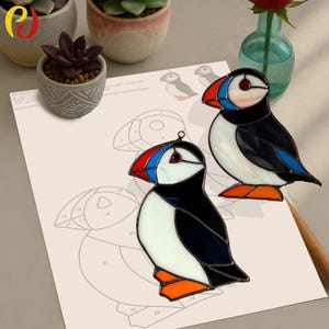 May include: Two stained glass puffin ornaments. The birds have black, white, red, blue, and orange details. One is hanging, and the other is standing. A sheet of paper with a puffin outline is in the background.