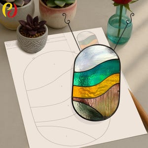 Peut inclure: Un panneau de vitrail avec un motif ondulé dans les tons de vert, jaune et marron. Le panneau est suspendu à un cadre en métal noir et est affiché sur un papier blanc avec un motif.