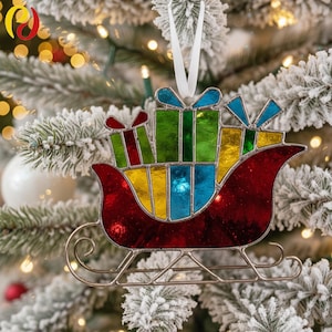 Puede incluir: Un adorno navideño de vidriera con un trineo rojo lleno de coloridos regalos. Los regalos son verdes, amarillos y azules con lazos de cinta blanca. El adorno cuelga de una cinta blanca sobre un árbol de Navidad nevado.