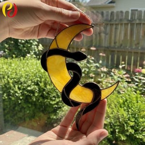 May include: A stained glass suncatcher with a yellow crescent moon and a black snake design. The suncatcher is suspended by a silver chain. The background shows green foliage and a glimpse of a building.
