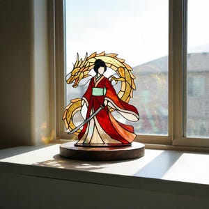 May include: Stained glass artwork of a woman in a red kimono with a sword, and a golden dragon. The piece is mounted on a round wooden base. The artwork is set against a window.