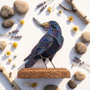 May include: A stained glass raven sculpture featuring iridescent dark blue and purple glass panels held together by dark metal solder lines. The raven holds a small silver-coloured metal knife in its beak and stands on a rectangular wooden base.