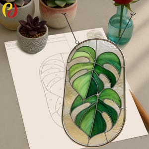 May include: Stained glass window art depicting two green monstera leaves. The leaves are in shades of green, set against a clear glass background with a silver metal frame. The art is suspended by a chain.