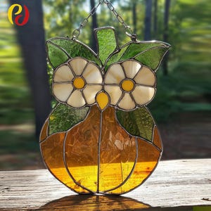 May include: Stained glass owl suncatcher with a chain for hanging. The owl has white and yellow flower-shaped eyes, green leaves, and a golden-orange body. The suncatcher is displayed outdoors on a wooden surface with a blurred green background.
