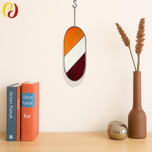 Puede incluir: Una ventana de vitral colgada en una pared blanca. La ventana tiene forma ovalada y presenta tres colores: naranja, blanco y burdeos. La ventana cuelga de una cadena negra.