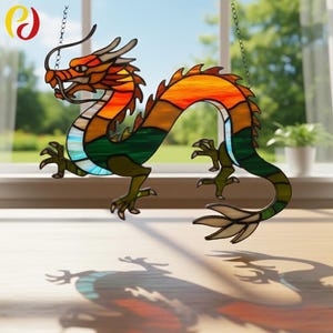 以下が含まれることがあります： カラフルなデザインのステンドグラスのドラゴンサンキャッチャー。ドラゴンはオレンジ、黄色、緑、青の色合いです。チェーンで吊り下げられており、下の表面に影を落としています。ドラゴンのデザインは複雑で詳細です。