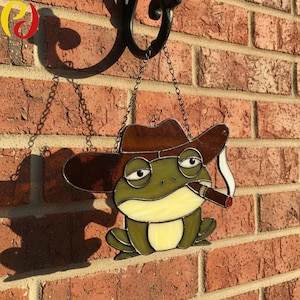 Puede incluir: Vidriera de una rana con sombrero de vaquero marrón y fumando un cigarro. La rana tiene cuerpo verde, vientre amarillo y cuelga de una cadena. El fondo es una pared de ladrillos rojos.