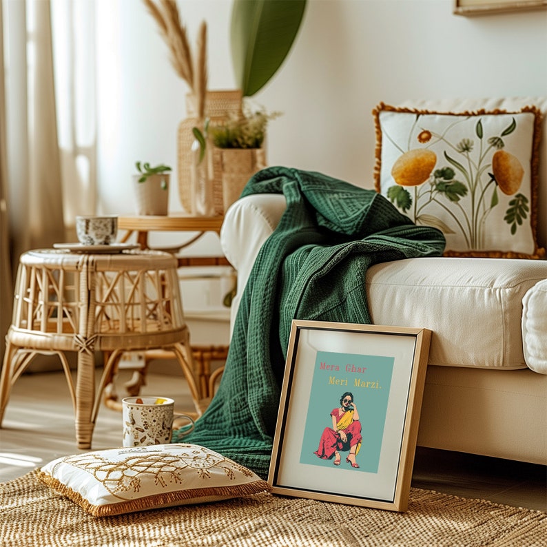 May include: A framed print with a colorful illustration of a woman in a pink and yellow outfit. The text on the print reads "Mera Ghar Meri Marzi". The print is sitting on a white couch with a green throw blanket and a floral pillow.