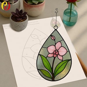May include: A stained glass teardrop-shaped ornament featuring a pink orchid with green leaves and a black outline. The ornament hangs from a decorative chain. A partially visible sheet of paper with a numbered diagram is in the background.