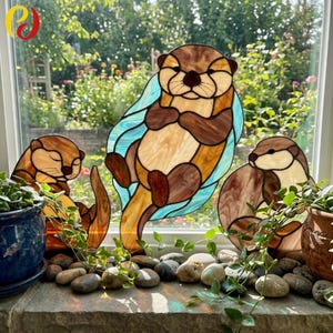 Pode incluir: Adesivos de janela de vidro manchado de lontra em tons de castanho, bege e azul. As lontras estão em várias poses, com uma flutuando de costas. Os adesivos de janela são exibidos em uma janela com plantas e pedras.