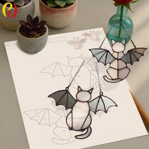 May include: Two stained glass cat ornaments with bat wings hang from chains. The cats are white and gray, with gray wings. A sheet of paper with a bat cat pattern and two small potted succulents are in the background.