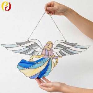 May include: Stained glass angel with outstretched wings, featuring a flowing blue and yellow dress. The angel's wings are a mix of white, gray, and blue. A silver chain is attached for hanging. The angel's head and arms are in shades of pink and yellow.