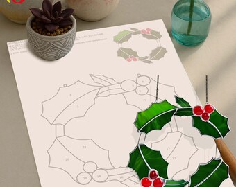 Op de afbeelding: Een groene en rode hulstkrans-ornament met een glas-in-lood-uiterlijk hangt voor een papieren sjabloon. De sjabloon toont een lijntekening van de krans. Kleine vetplanten in pot en een vaas met een rode roos staan op de achtergrond.