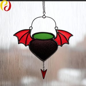 Könnte beinhalten: Ein Glasfenster-Suncatcher mit einem Kesseldesign mit roten Fledermausflügeln und einer grünen Flüssigkeit. Der Kessel ist dunkelviolett mit schwarzem Umriss und einem roten Pfeilanhänger. Er hängt an einer schwarzen Metallkette.