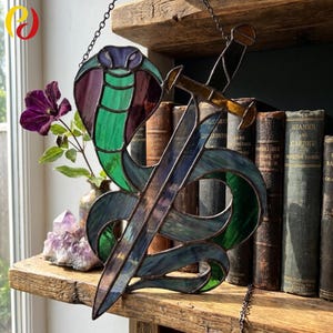 Op de afbeelding: Gebrandschilderd glaskunstwerk met een cobra en een zwaard. De cobra is in groen, paars en blauw, het zwaard heeft een gouden handvat. Het kunstwerk hangt voor een plank met oude boeken.