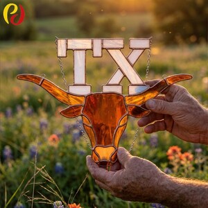 May include: A stained glass Texas Longhorn with the letters "TX" at the top. The Longhorn is amber, with white and clear glass letters. The piece is held by hands against a blurred floral background.