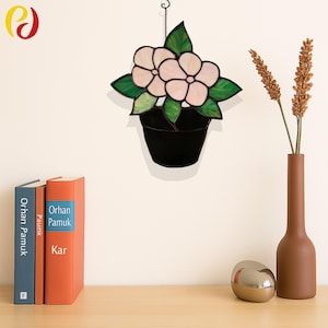 Puede incluir: Obra de arte floral en vidriera con dos flores de color rosa pálido con hojas verdes en una maceta negra. La obra cuelga de una cadena contra una pared color crema. Un jarrón marrón con trigo y libros están sobre una superficie de madera.