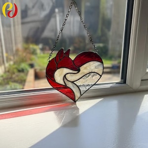 May include: Stained glass heart-shaped suncatcher featuring a red fox design. The fox is depicted in red and white glass, set against a clear and textured background. The suncatcher hangs from a metal chain, perfect for window or wall decor.