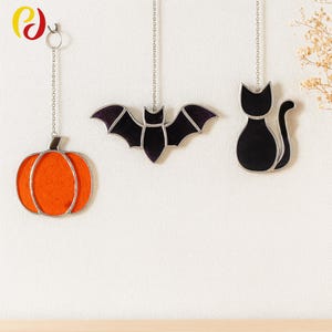 Puede incluir: Tres decoraciones colgantes de Halloween: una calabaza naranja, un murciélago negro con detalles morados y un gato negro. Cada pieza está delineada en plata y cuelga de una cadena. La calabaza tiene una forma clásica.