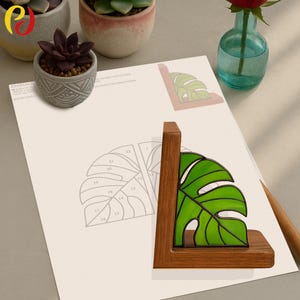 May include: A wooden bookend featuring a vibrant green stained glass monstera leaf design. The bookend is made of dark wood and sits on a light-colored surface. A pencil and a vase with a red rose are also visible.