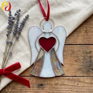 May include: Stained glass angel ornament with a red heart center and white wings. The angel has a silver metal outline and a red ribbon for hanging. The ornament is displayed on a wooden surface.