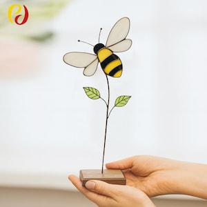 May include: A stained glass bee on a wire stem with green leaves, mounted on a small wooden base. The bee has yellow and black stripes, with white wings and a black head. The artwork is held in a person's hands.